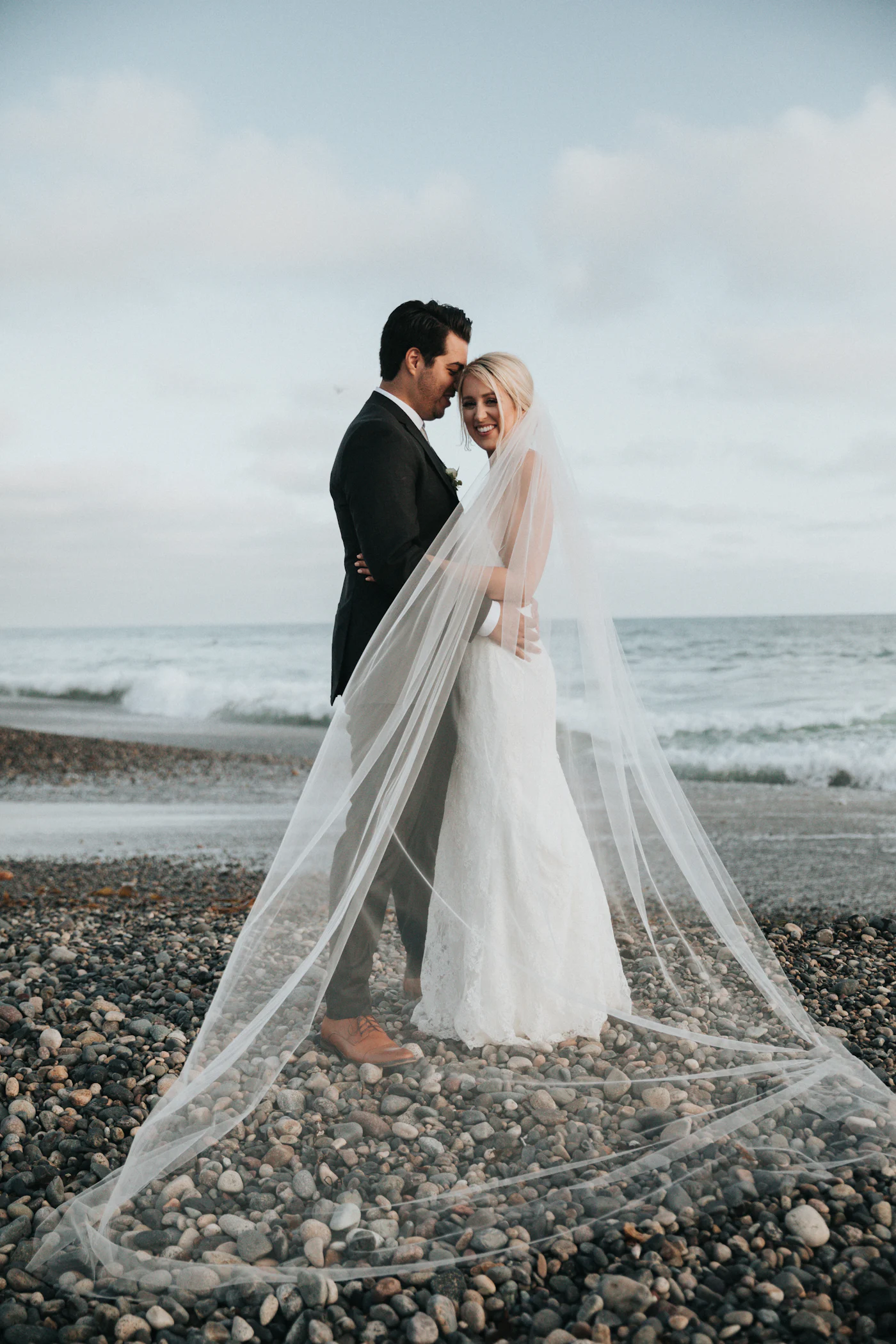 Couple embracing on a rocky Pacific coast beach, bride's cathedral veil sweeping in the ocean wind